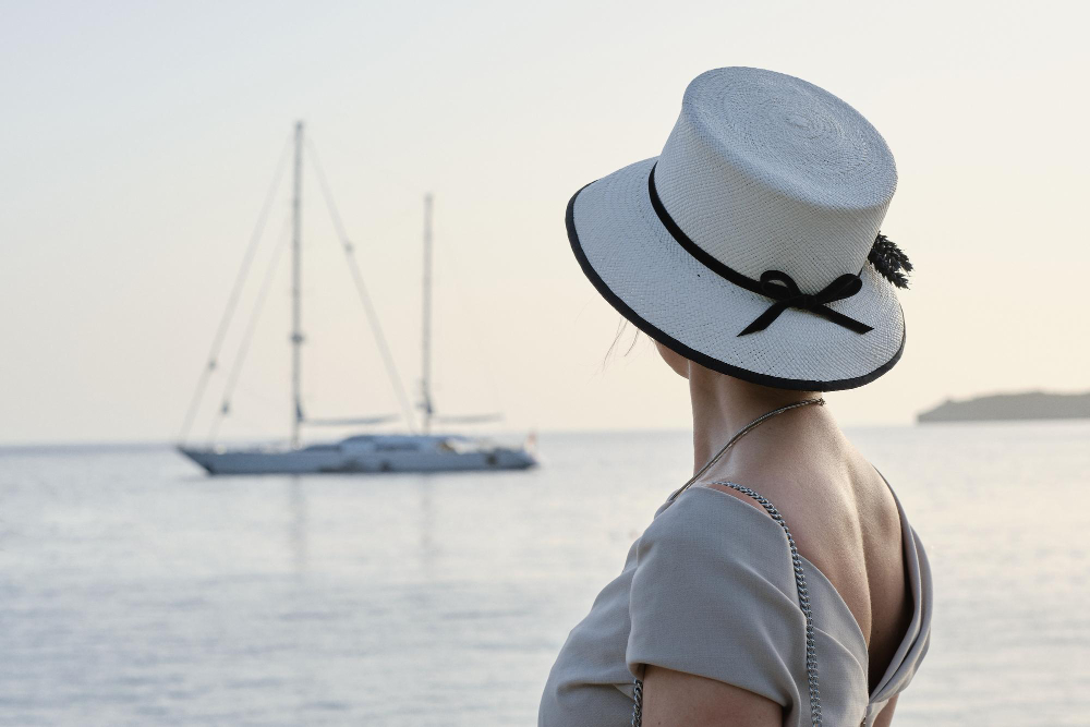 back-view-of-woman-with-hat-standing-in-dress-looking-out-towards-yacht-in-blue-sea-and-sky